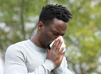 A man is sneezing into a tissue while standing outdoors.