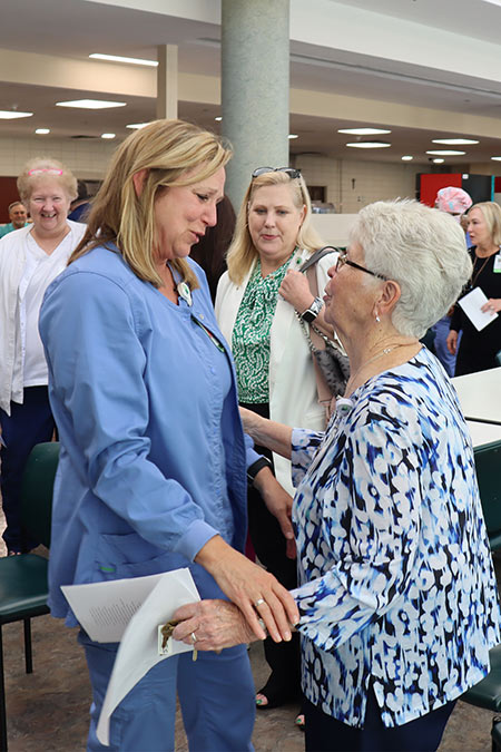 Alison White woman in medical scrubs warmly greets an elderly woman in a patterned blouse, holding her hand and smiling, while a group of people watches in the background.