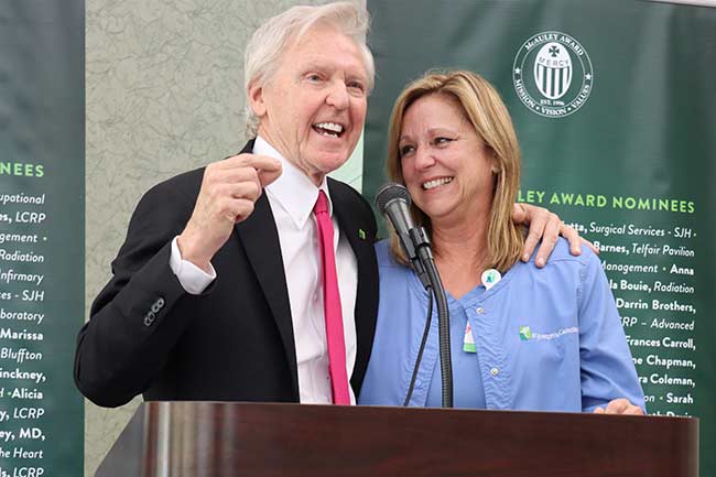 St. Joseph’s/Candler President & CEO Paul P. Hinchey is announcing Alison White as the recipient of the 30th Annual McAuley Award at a podium, while a smiling Alison White in medical scrubs stands beside him.